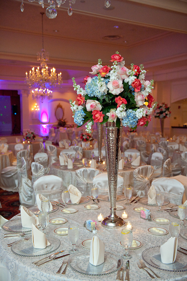 reception seating decoration - dark pink, light pink, light blue, light green, and white floral arrangement in tall silver vase as centerpiece, white table cloths with white chair covers and napkins, ivory candles on table tops, and crystal chandeliers - photo by Houston based wedding photographer Adam Nyholt